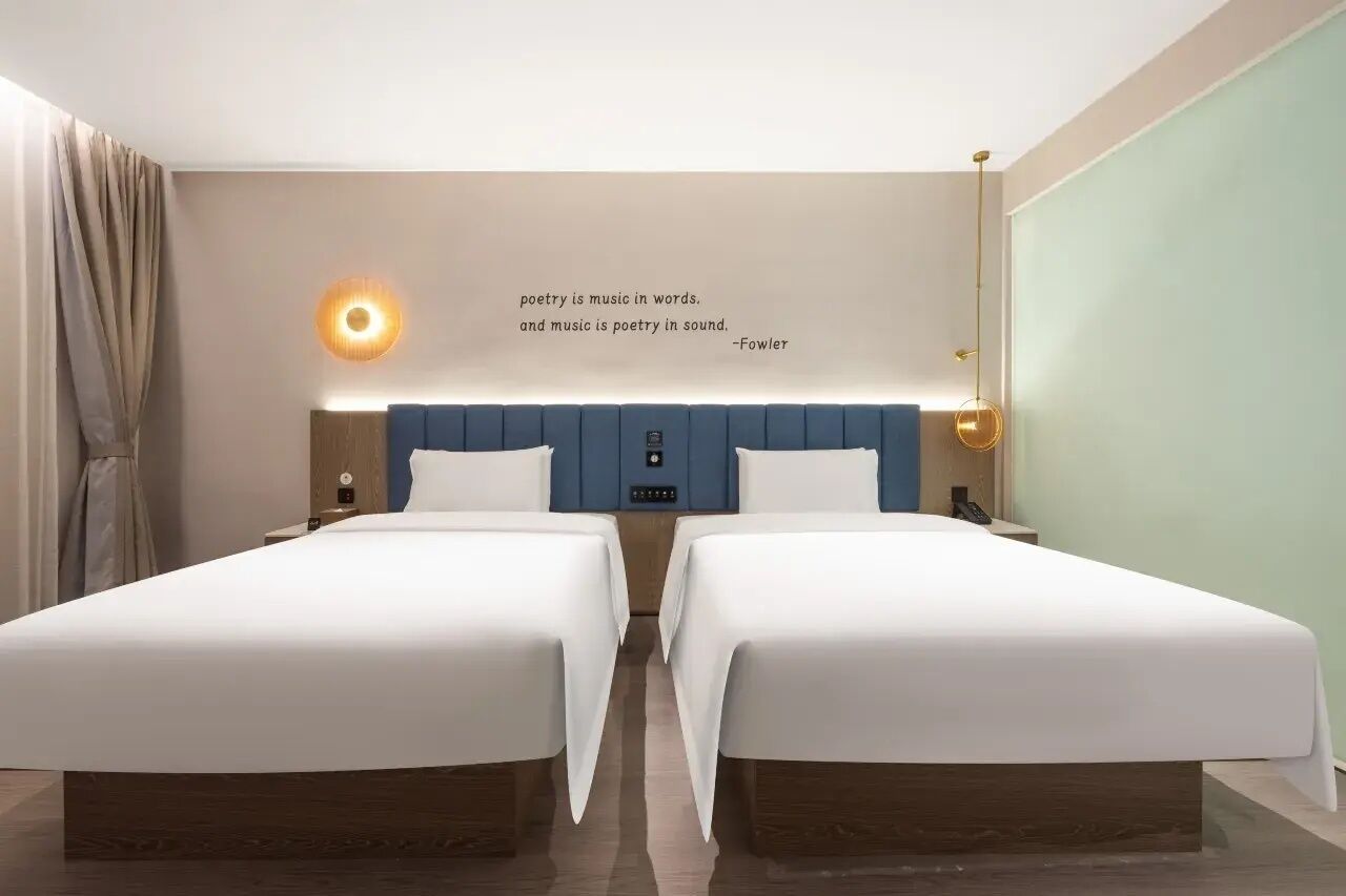 Une chambre d’hôtel avec deux lits, une lampe fixée au mur et une citation de Fowler.