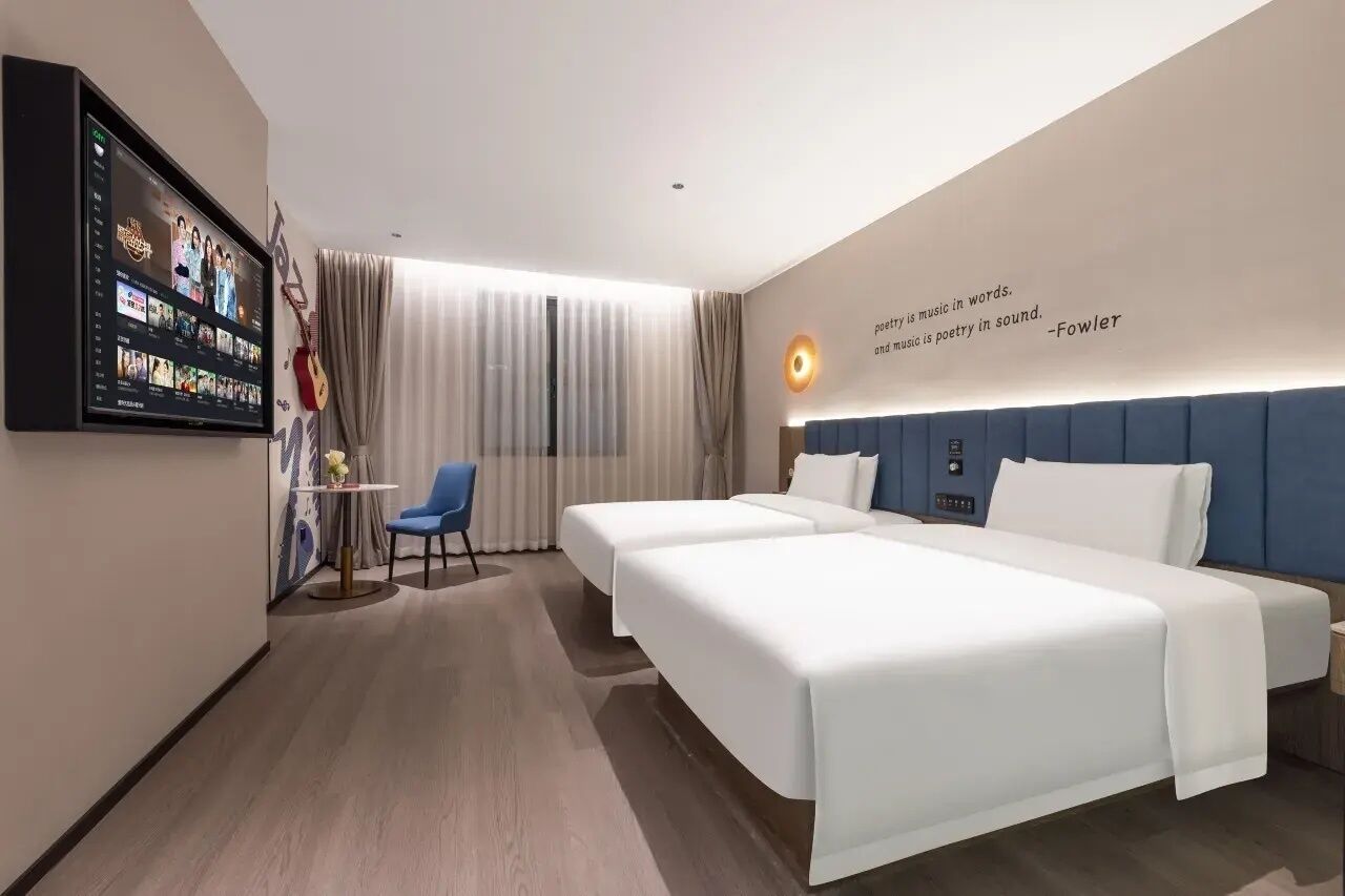 A modern hotel room with two beds, a TV mounted on the wall, a desk with a chair, and a quote on the wall.