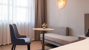 A modern hotel room with a grey sofa, a blue chair, a small round table with a vase of flowers, and a wall-mounted light fixture. - MANNSIC Hotel- Shenzhen Haishang Shijie (Shenzhen)
