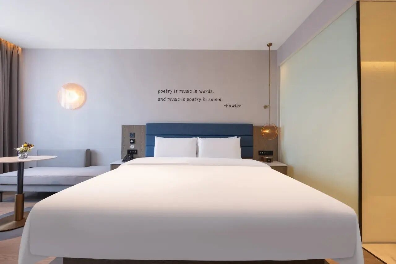 A modern hotel room with a large bed, a bedside table, a small table with flowers, a wall-mounted light, and a quote about poetry and music.