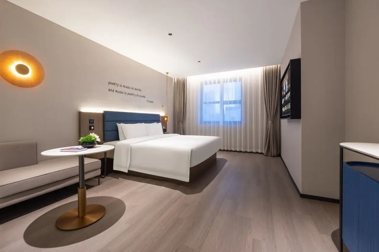 A modern hotel room with a bed, a sofa, a small table, and a wall-mounted TV.