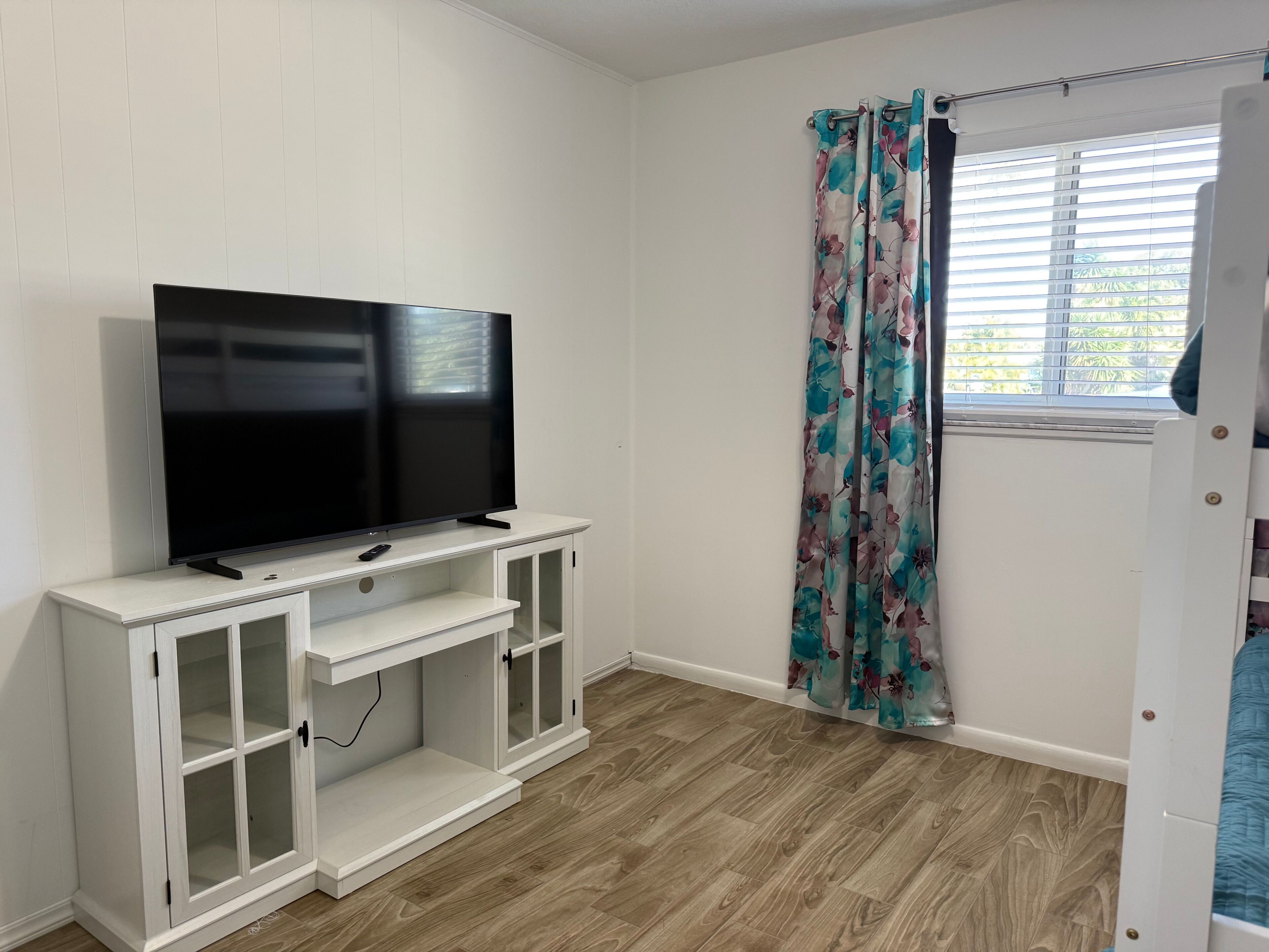 A room with a flat-screen TV on a white entertainment unit, a window with blinds, and a door with a blue curtain.