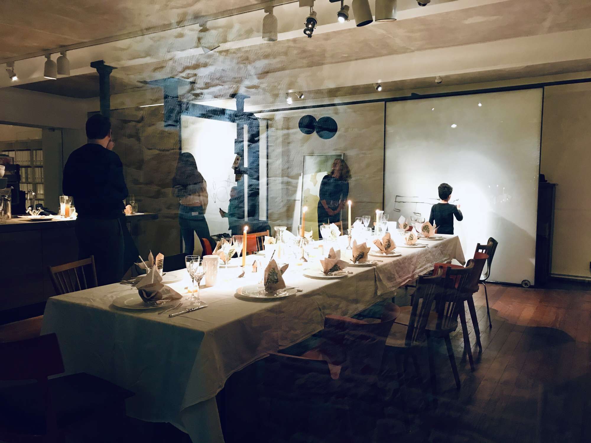 Une longue table à manger, décorée de bougies et de assiettes, se trouve dans une pièce dont un mur est en pierre et qui est éclairée par des luminaires modernes.