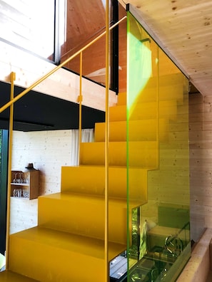 Una escalera con peldaños amarillos y barandilla de cristal, que baja a un nivel inferior.