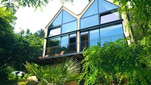 A modern house with large glass windows, a balcony with chairs, and lush greenery around. - Burgstrasse Apartment West with garden and sauna (Altleiningen)