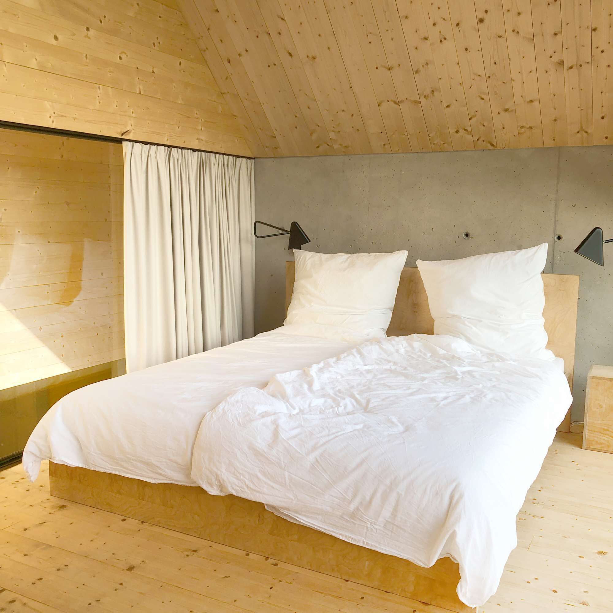 Dormitorio de una cabaña de madera con cama, cortinas blancas y una pared de concreto.