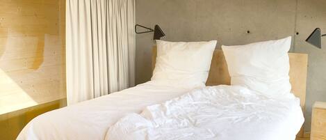 Dormitorio de una cabaña de madera con cama, cortinas blancas y una pared de concreto.