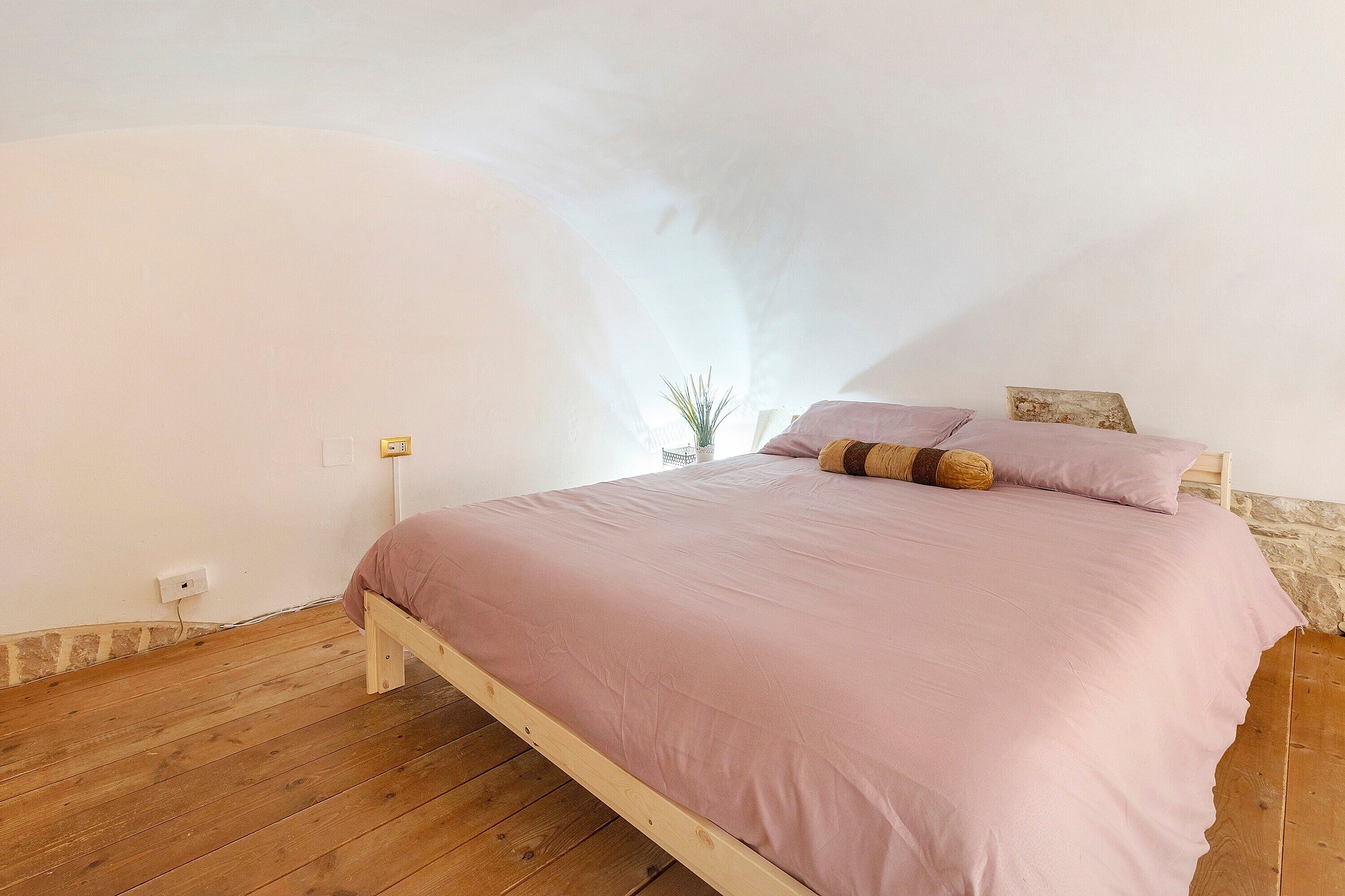 A bedroom with a wooden floor, a bed with a pink bedspread, and a stone wall.