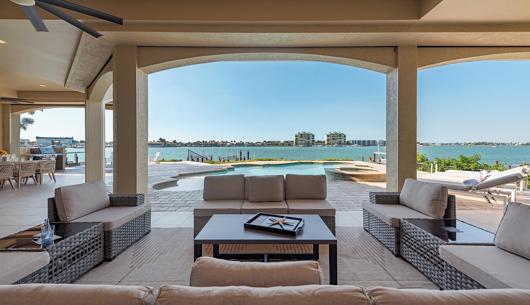 A covered outdoor seating area with a pool, lounge chairs, and a view of the water.