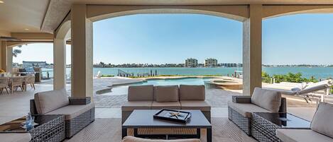 A covered outdoor seating area with a pool, lounge chairs, and a view of the water.