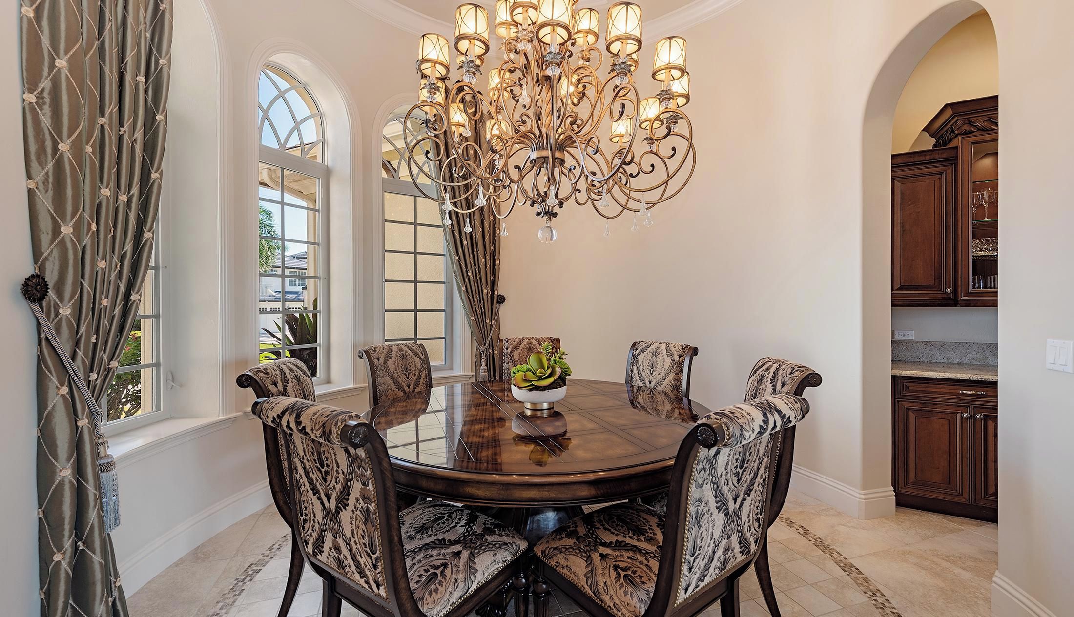 A dining room with a round table, six patterned chairs, a chandelier, and a view to the kitchen.