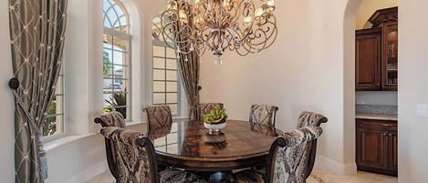 A dining room with a round table, six patterned chairs, a chandelier, and a view to the kitchen.