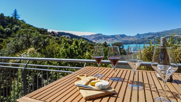 A wooden table with a cheese board, two wine glasses, and a bottle of wine on a balcony overlooking a scenic landscape with mountains and a lake. - Harbour Crest Retreat - Lyttelton Holiday Home (Lyttelton)