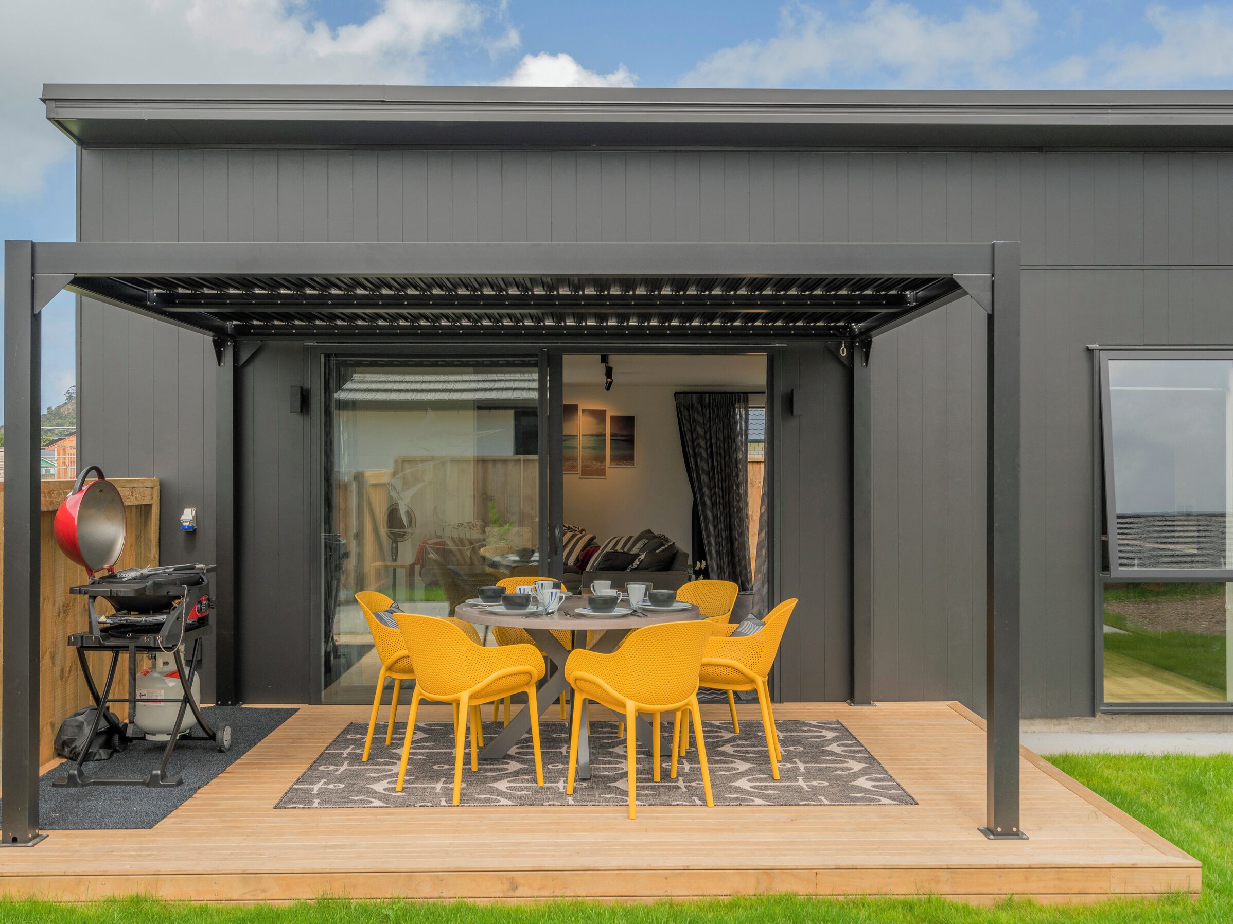 Un espace repas extérieur moderne, doté d’un patio couvert, d’un barbecue et offrant une vue sur l’intérieur grâce à une porte vitrée.