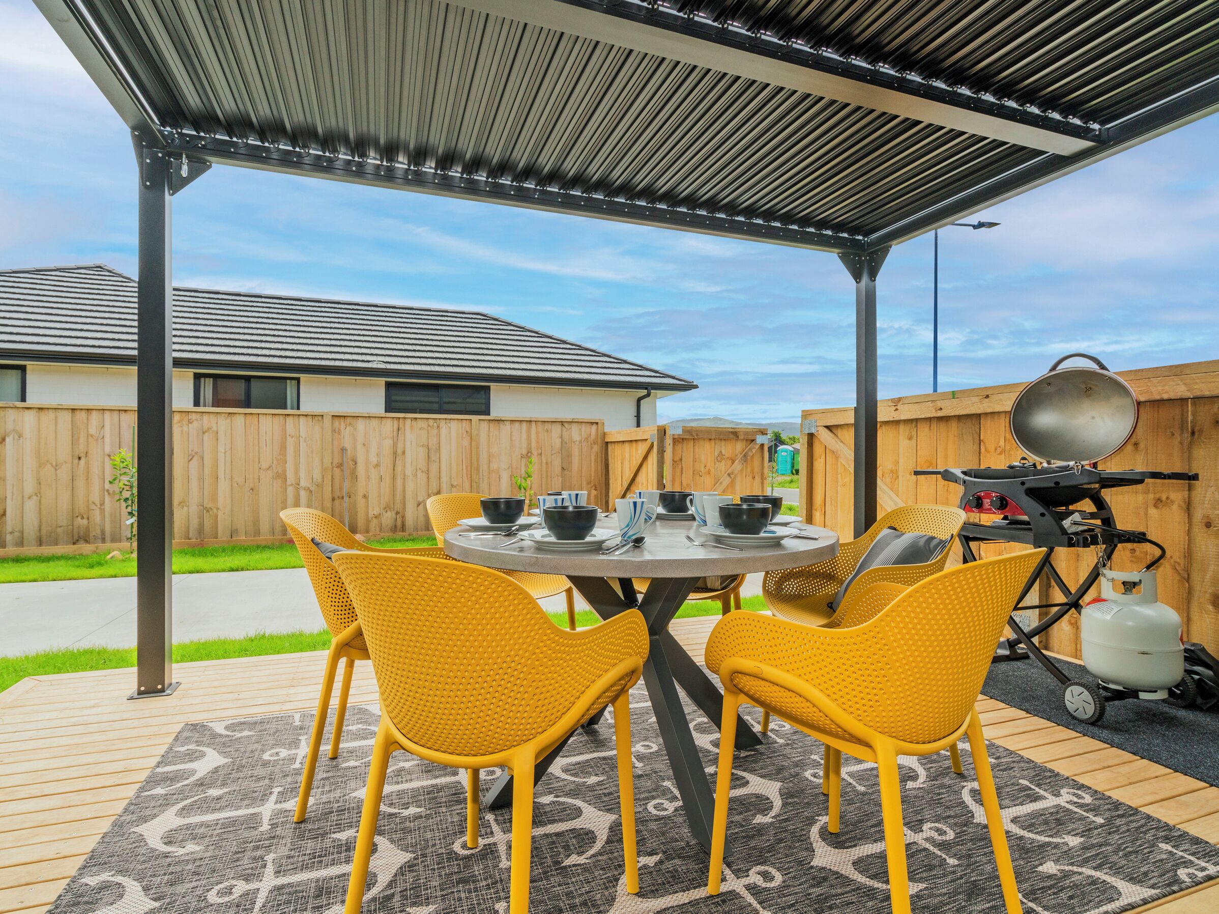 Een overdekt terras met een ronde tafel, gele stoelen, een barbecue en een draagbaar toilet.