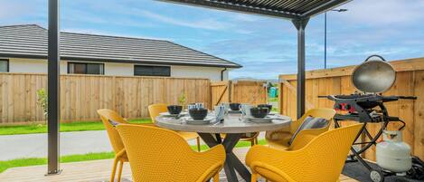 Een overdekt terras met een ronde tafel, gele stoelen, een barbecue en een draagbaar toilet.