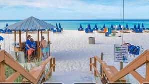 A beach with wooden walkways, a shaded area with a table and chairs, and a sign indicating beach services. - Splash! Kids Aqua park, Free rental chairs! (Panama City Beach)