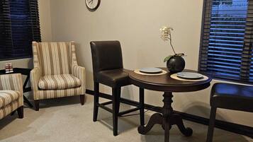 A dining area with a round table, two chairs, a clock on the wall, and a window with blinds.