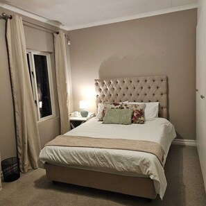 Ein Schlafzimmer mit einer gepolsterten Kopfteile, einem Bett mit einer beigefarbenen Bettdecke, einem Nachttisch mit Lampe und einem Fenster mit Vorhängen.
