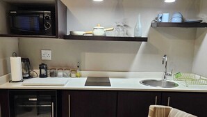 Eine kompakte Küche mit Mikrowelle, Kaffeemaschine und kleinem Kühlschrank. Zwei Pendelleuchten sind über der Arbeitsplatte angebracht, und ein Stuhl befindet sich im Vordergrund.