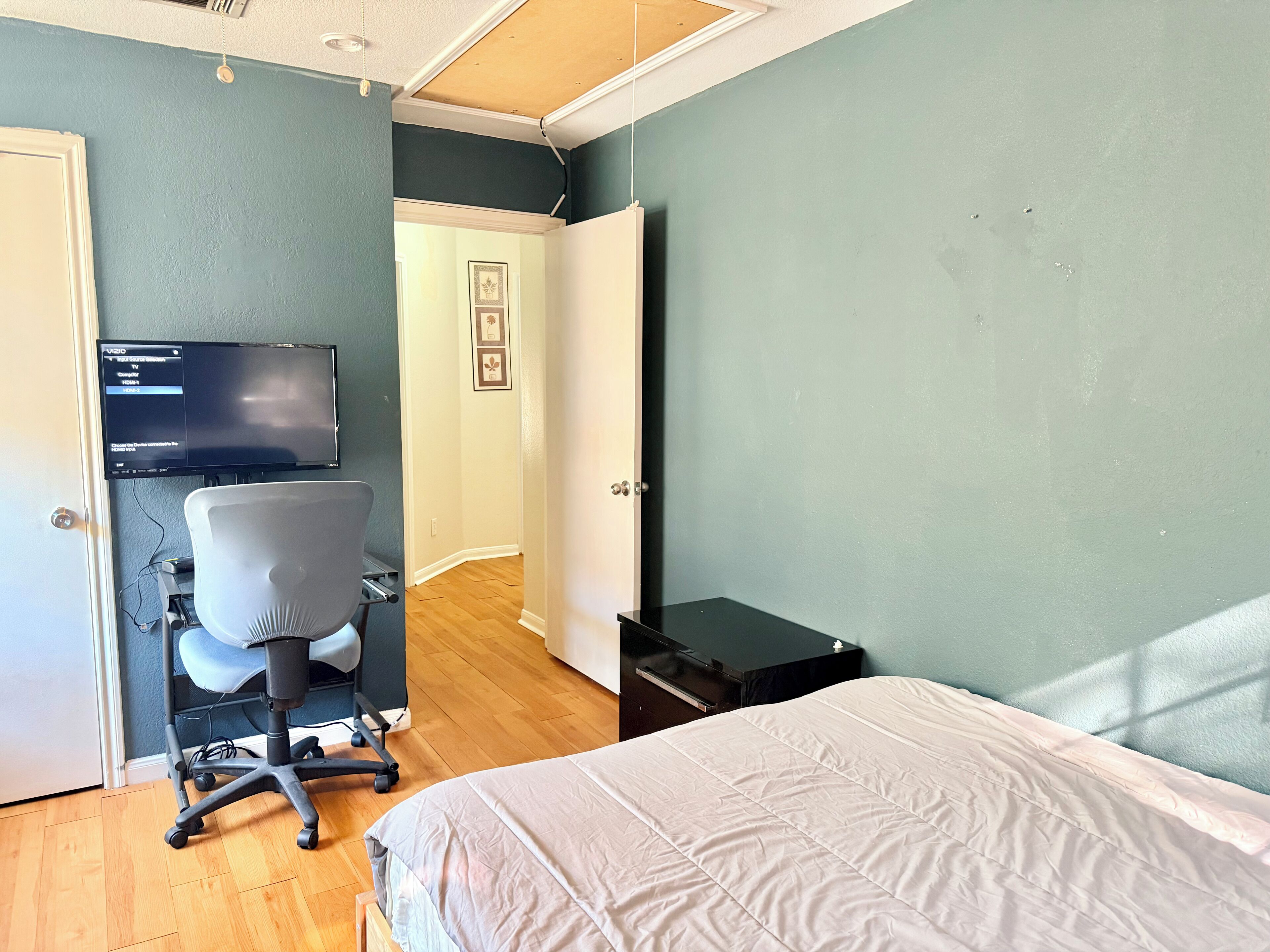 A bedroom with a bed, a desk with a chair, a TV on the wall, and a door leading to another room.