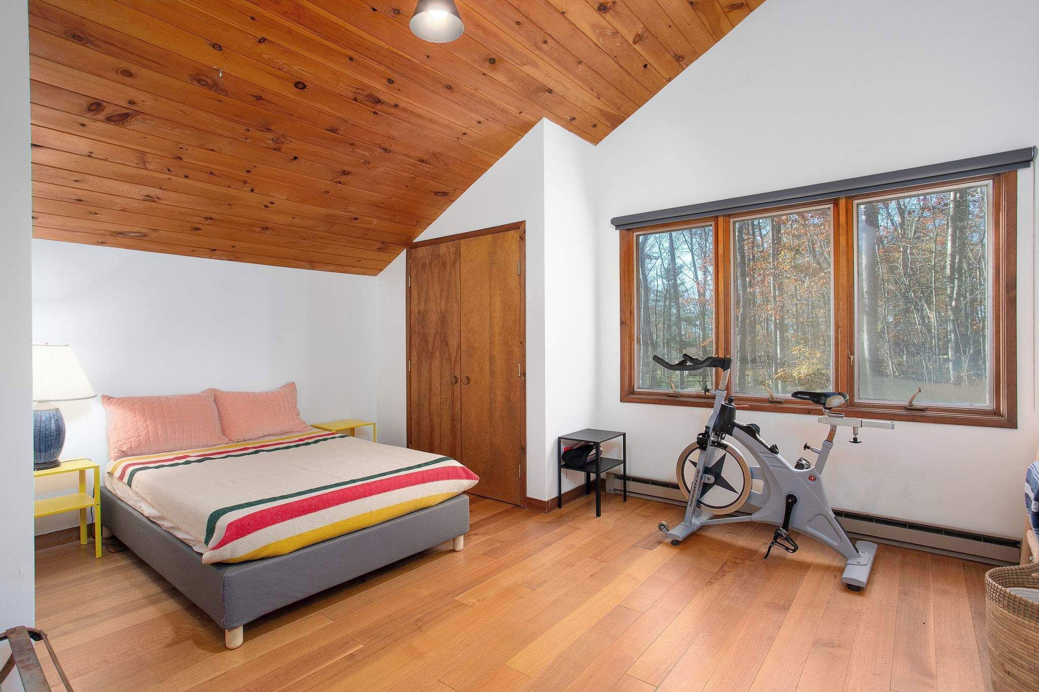 Een slaapkamer met een bed, houten plafond, raam met uitzicht op bomen en een hometrainer.