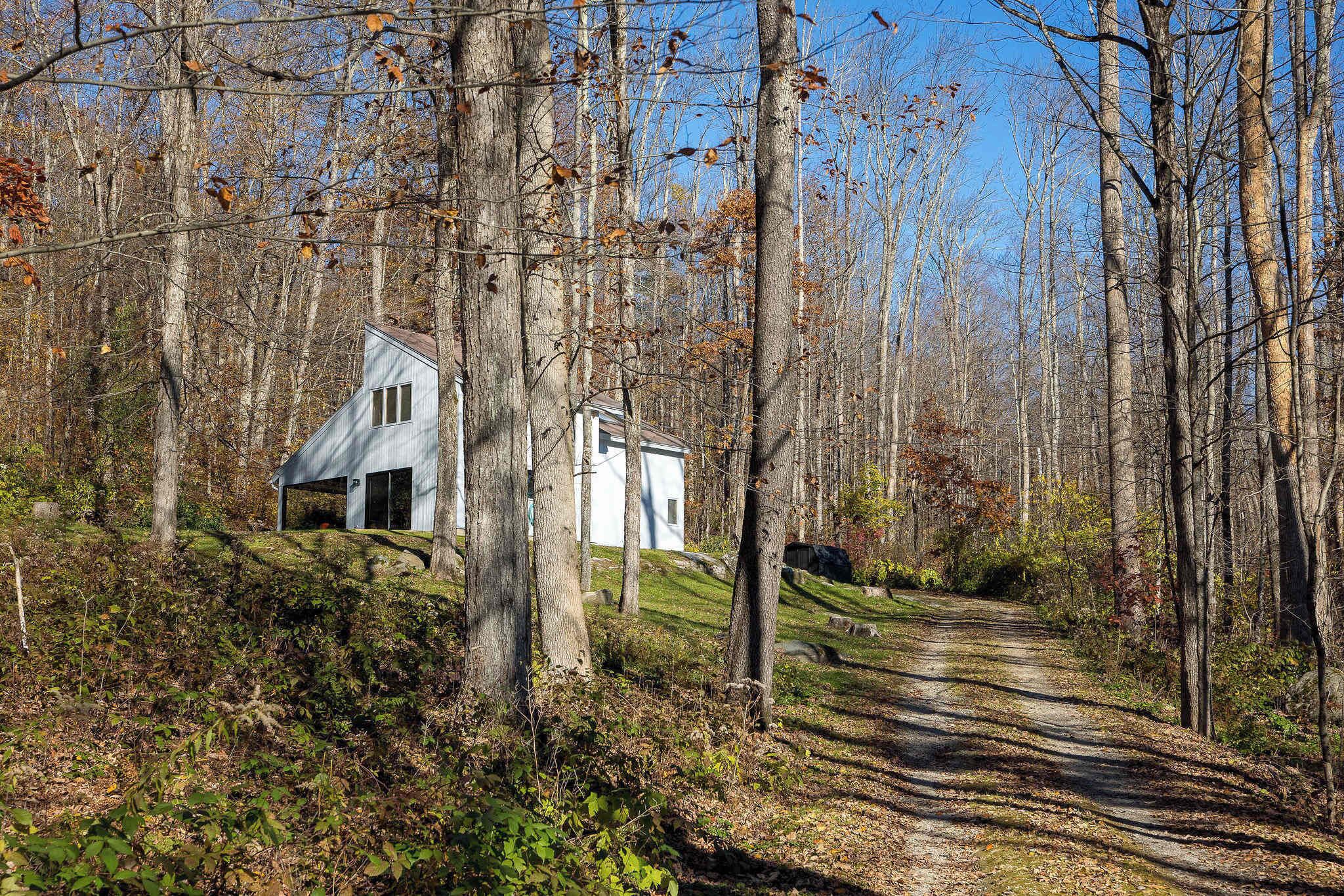 A rustic cabin with a sloped roof and a small porch, surrounded by a forested area with a dirt path leading to it.