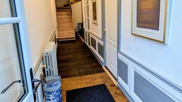 A narrow hallway with a wooden floor, white walls, and a black carpet. A radiator and a blue and white vase are visible on the left.