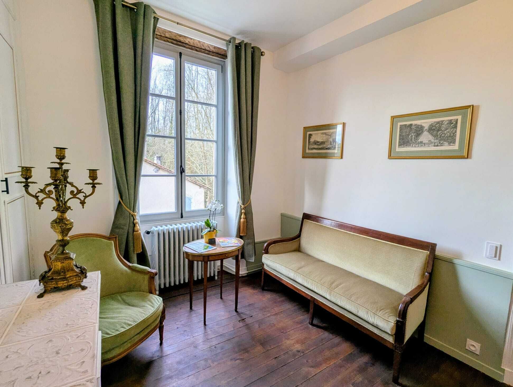 A room with a wooden floor, a green armchair, a sofa, a table with a plant, and two framed pictures on the wall.