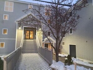 Edificio de dos pisos con una escalera de madera que lleva a una puerta principal, rodeado de nieve.