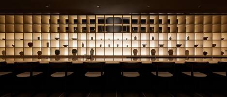 Un interior moderno con una larga barra y varias sillas, destacando una pared de estanterías iluminadas que exhiben diversos objetos.