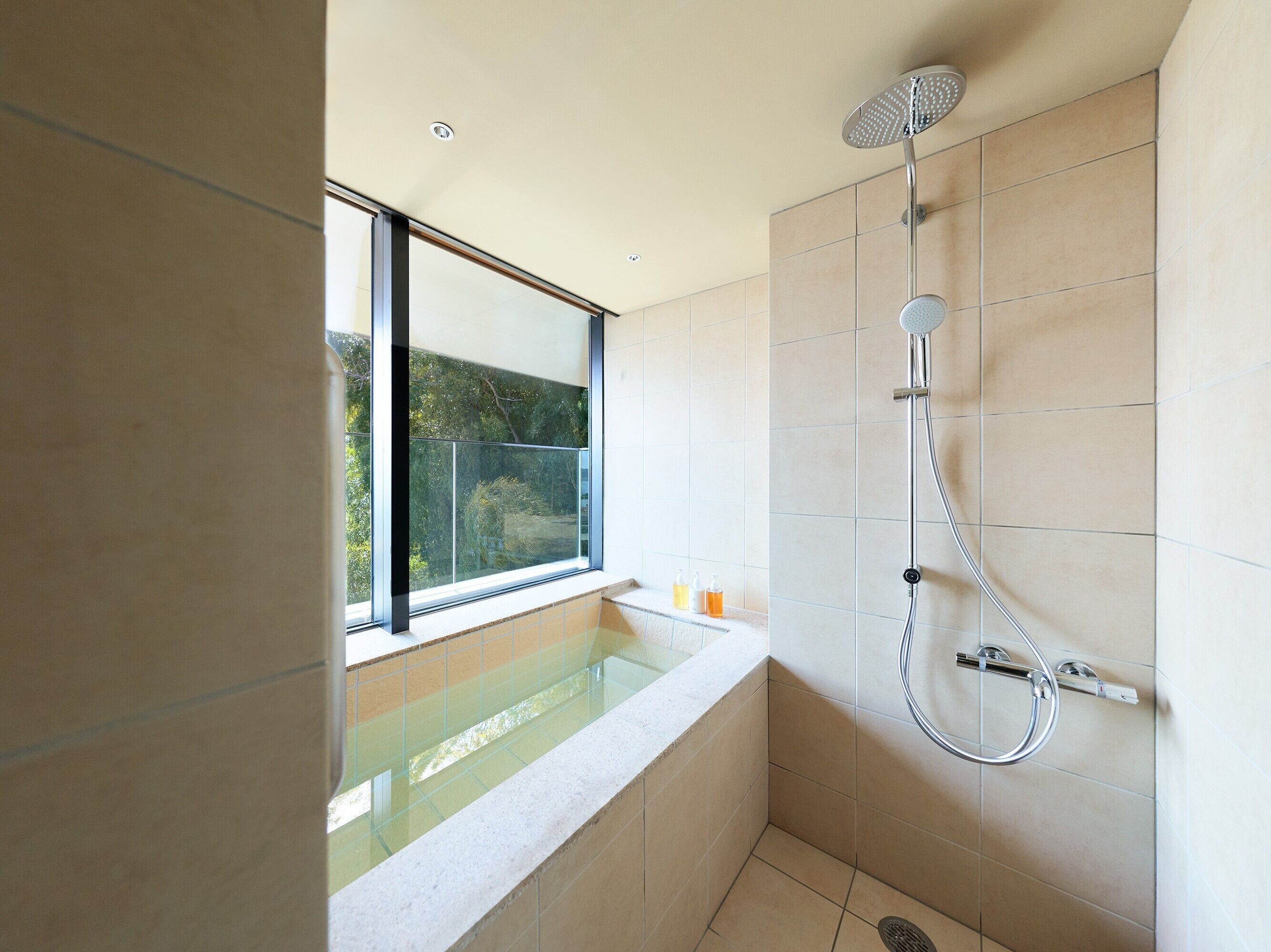 A modern bathroom with a glass-enclosed shower, a bathtub, and a window providing a view of greenery.