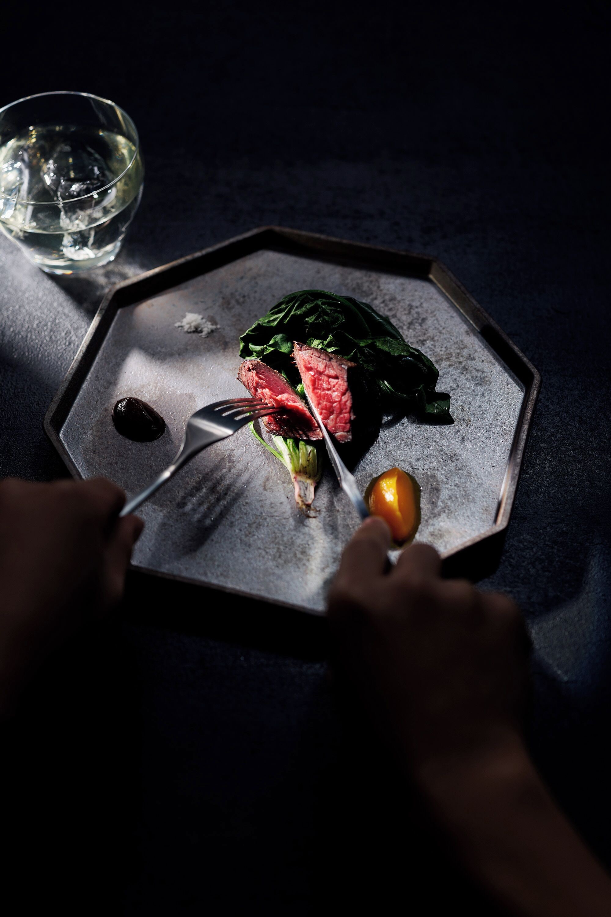 A person using a fork to eat a piece of meat with vegetables on a metal plate.