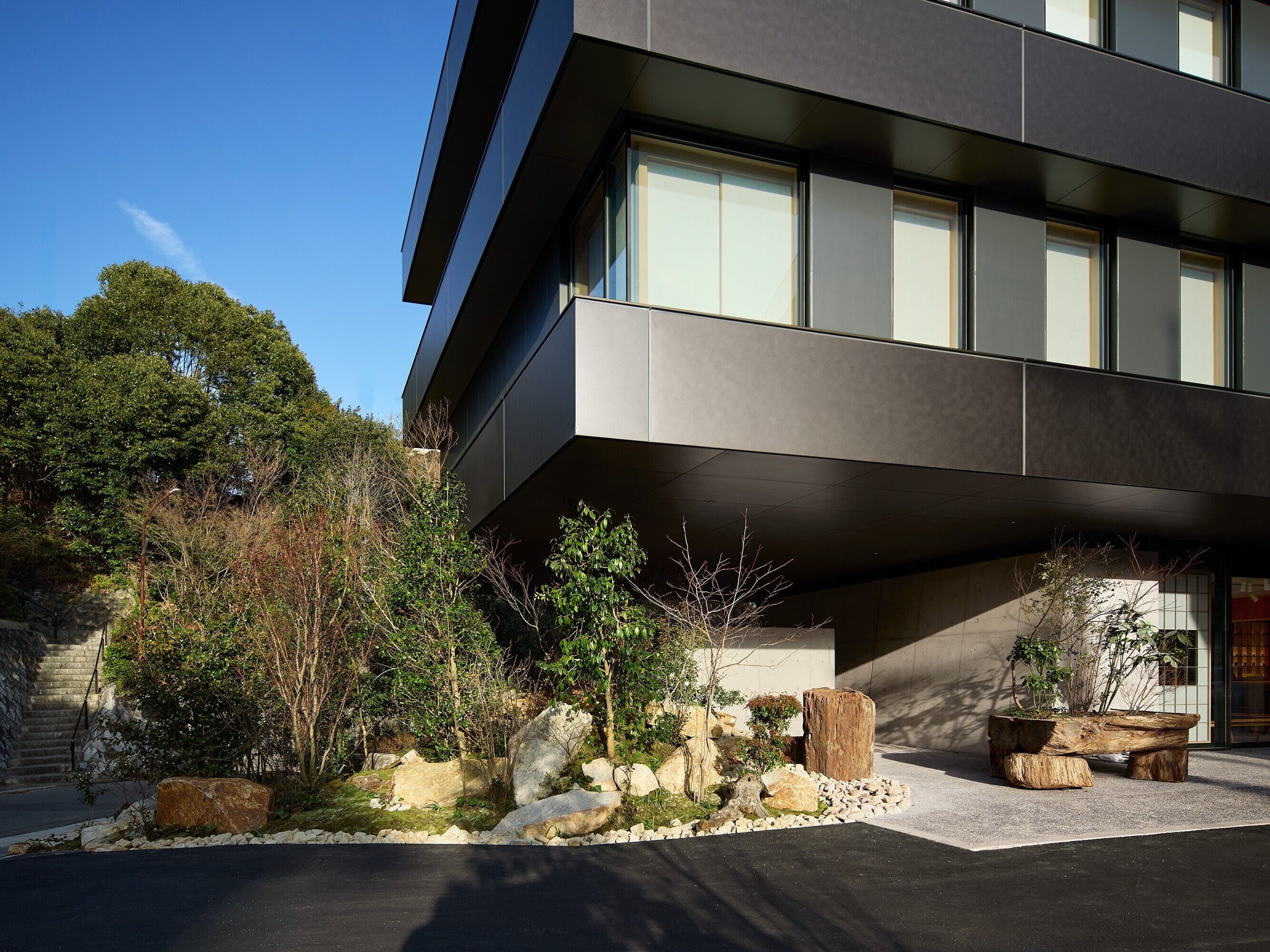 Un edificio moderno con grandes ventanales, una escalera de piedra y un jardín ajardinado con rocas y árboles.
