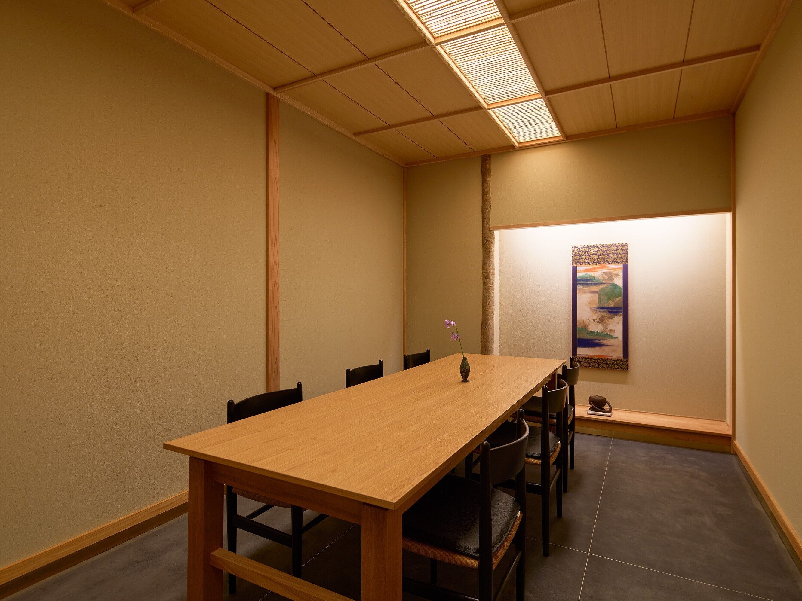A conference room with a long wooden table, black chairs, a vase with a flower, and a painting on the wall.