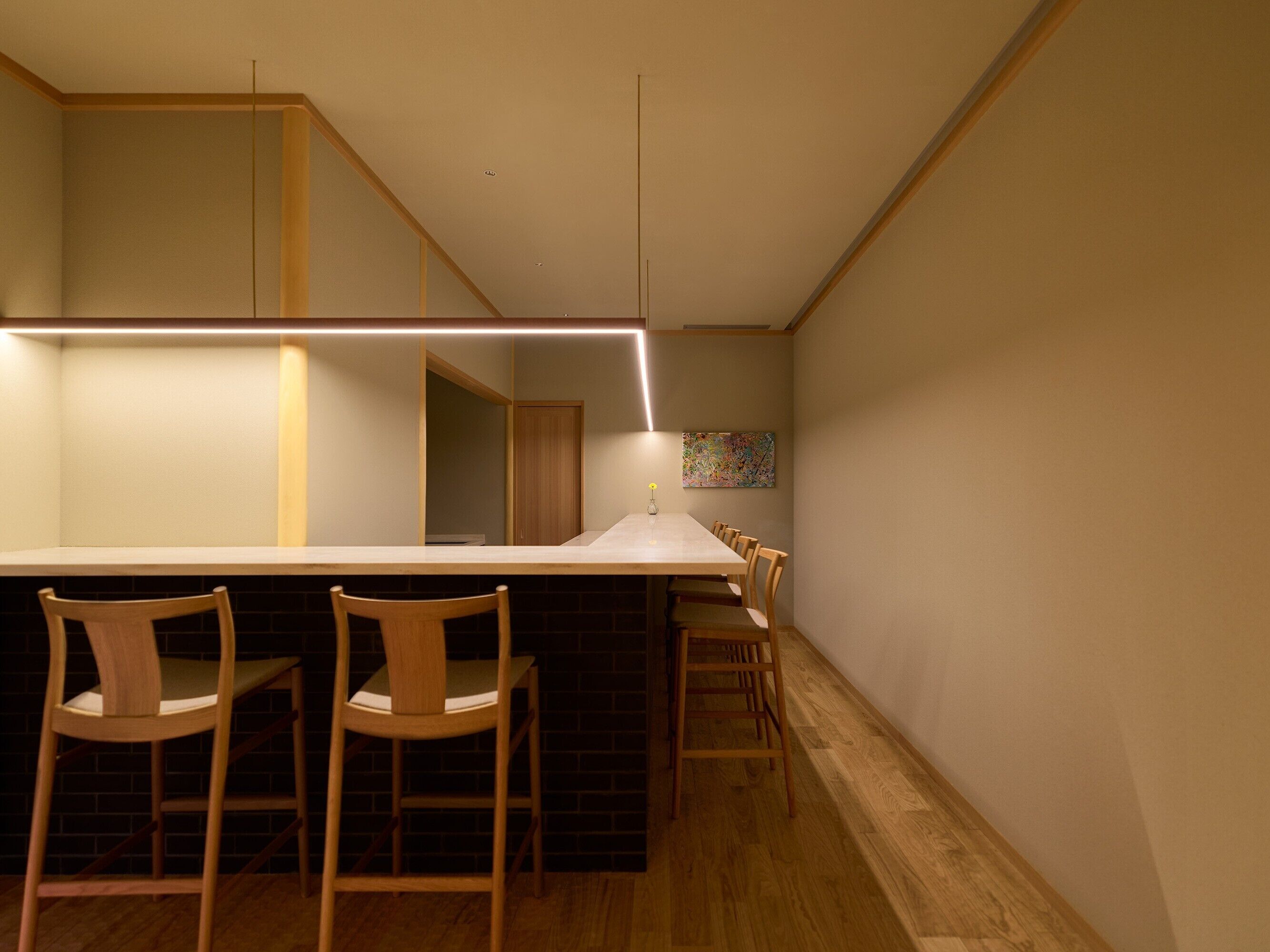A modern kitchen with a long countertop, wooden chairs, and a hanging light fixture.