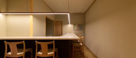A modern kitchen with a long countertop, wooden chairs, and a hanging light fixture.