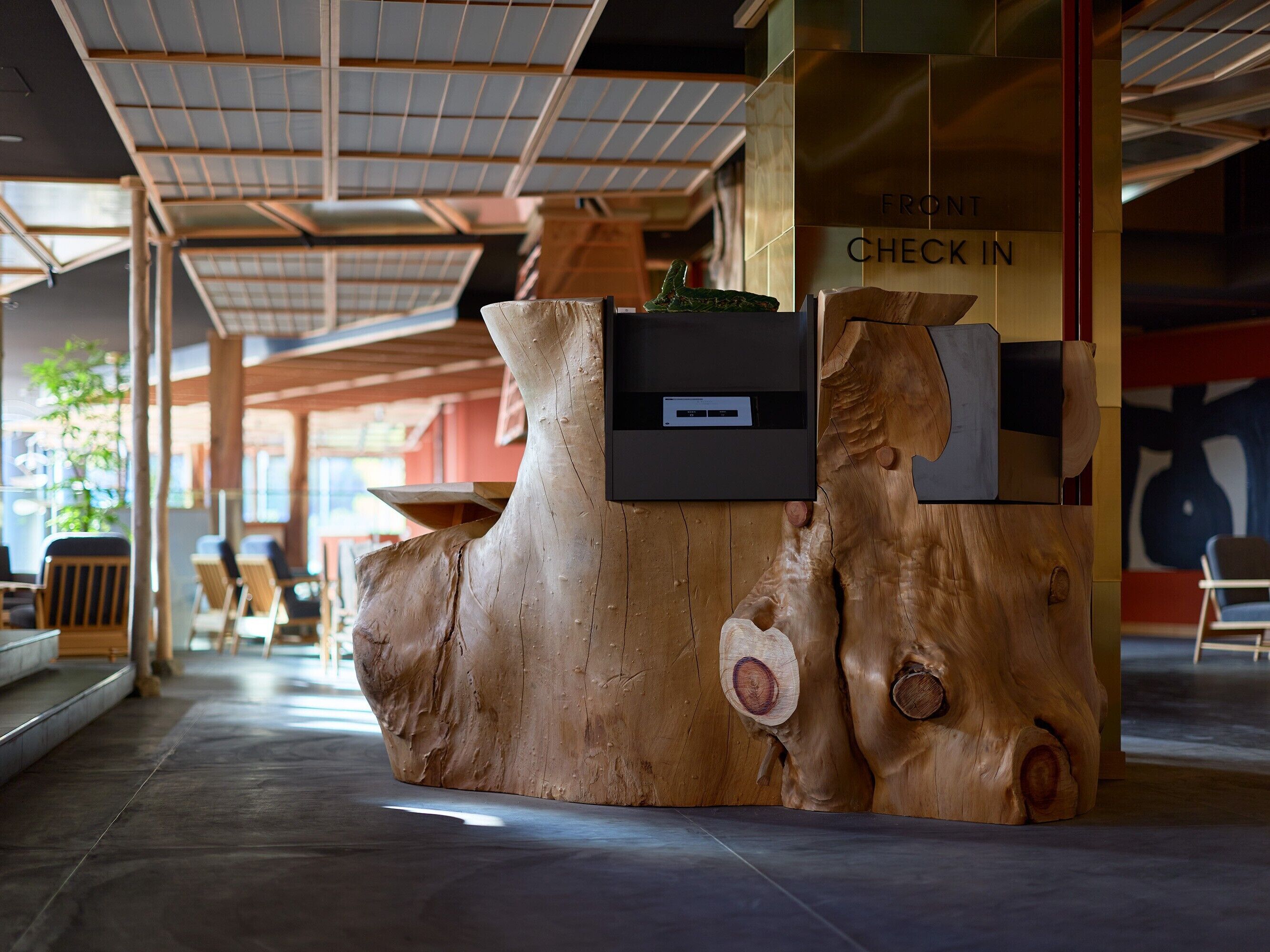 A reception desk made from a large tree trunk with a 'Check In' sign, surrounded by wooden chairs and tables, under a high ceiling with skylights.
