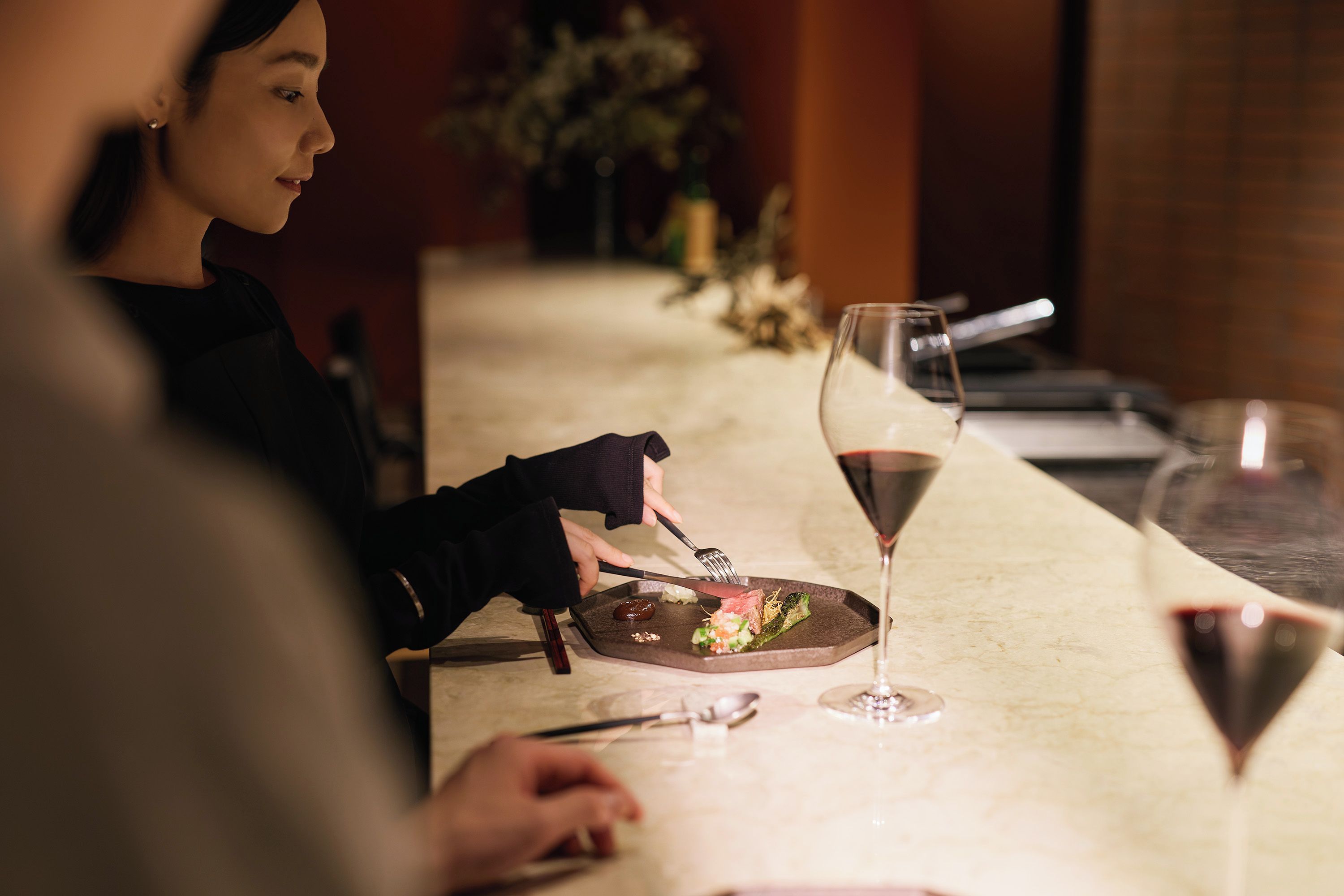 A dining table with a plate of food, a glass of red wine, and a person using cutlery.