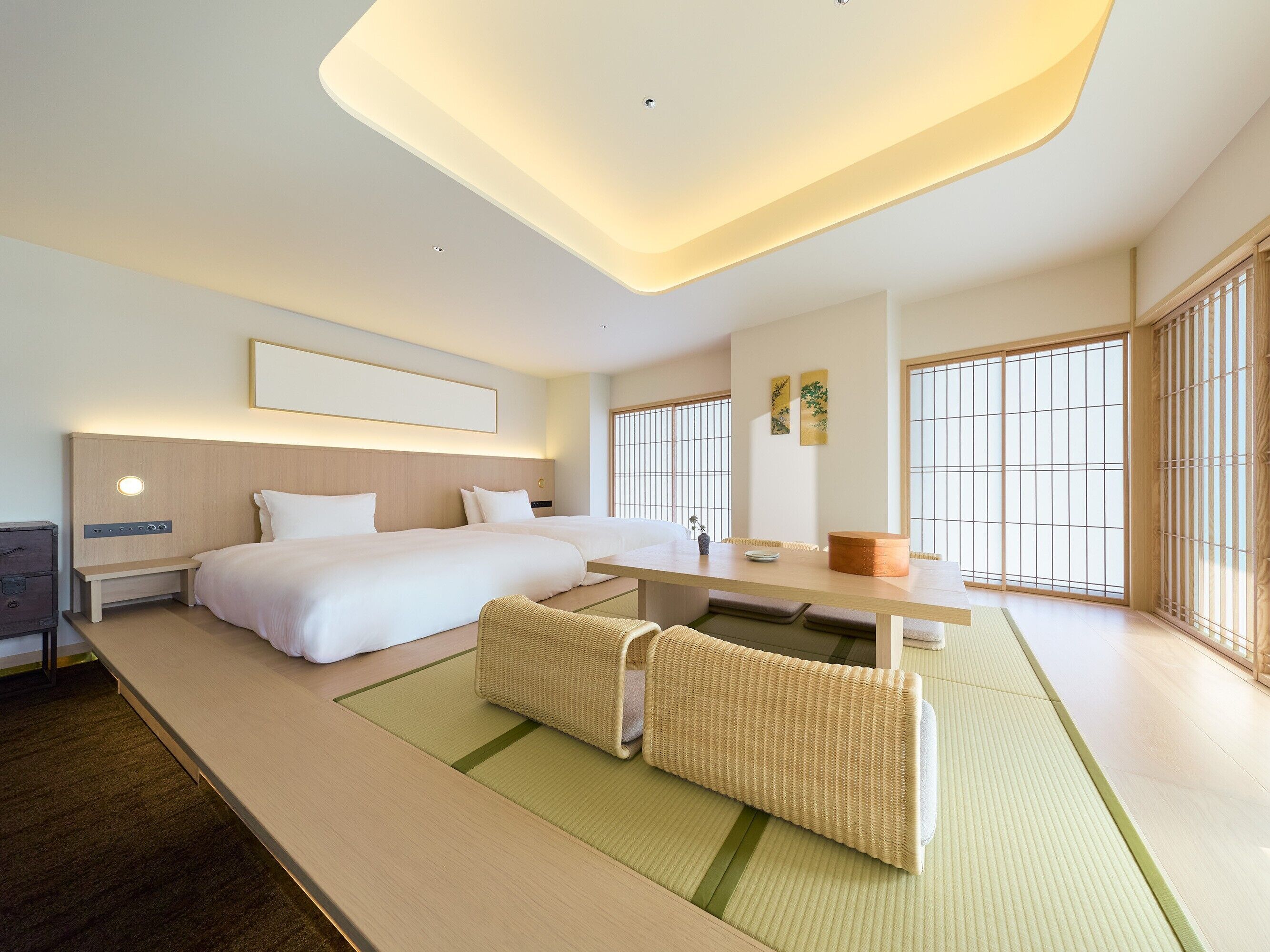 A modern hotel room with a large bed, a bedside table, a small desk, and a view of the outside through large windows.