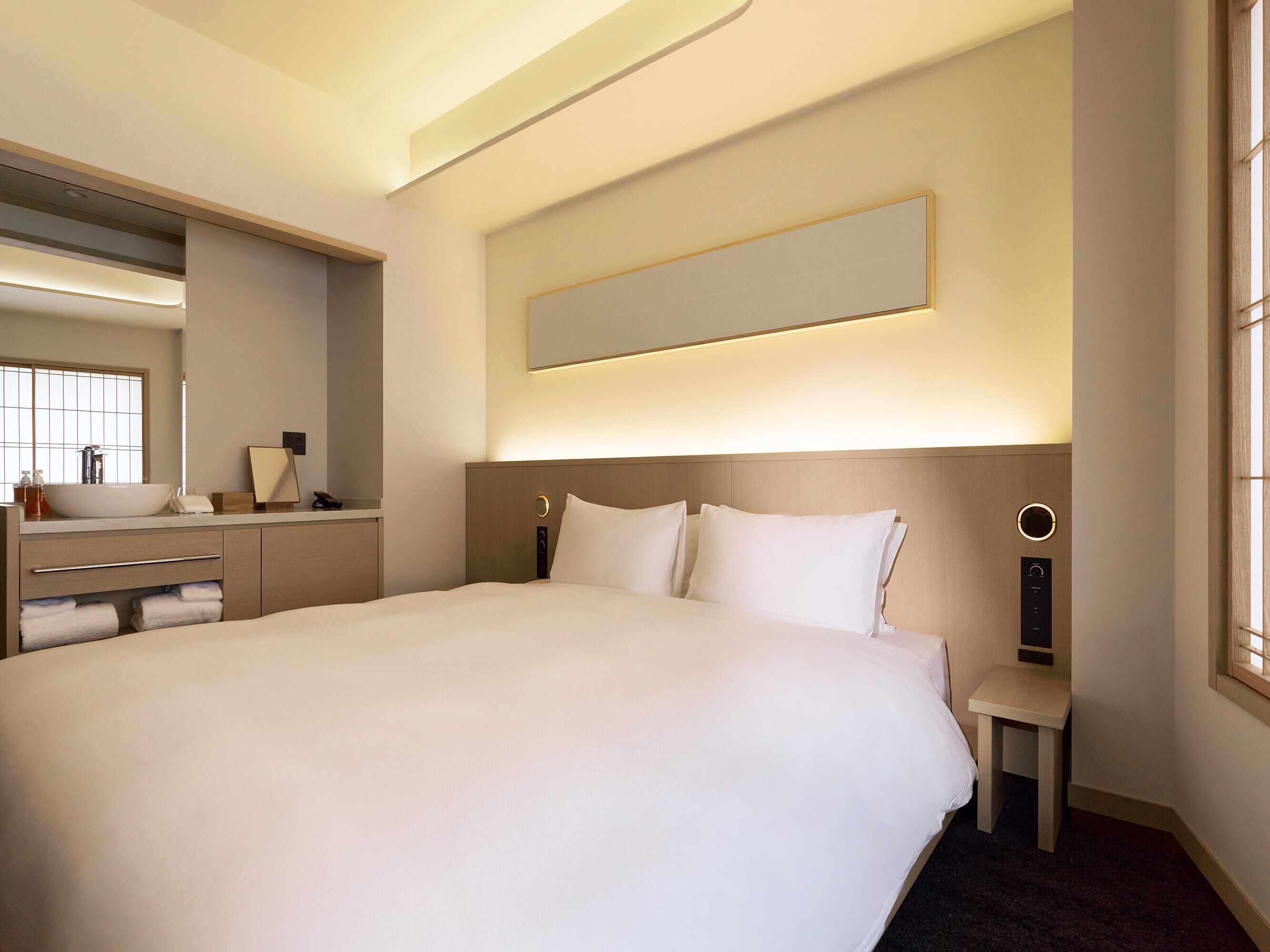 A hotel room with a large bed, a bedside table, a mirror, a vanity with a sink, and a window.