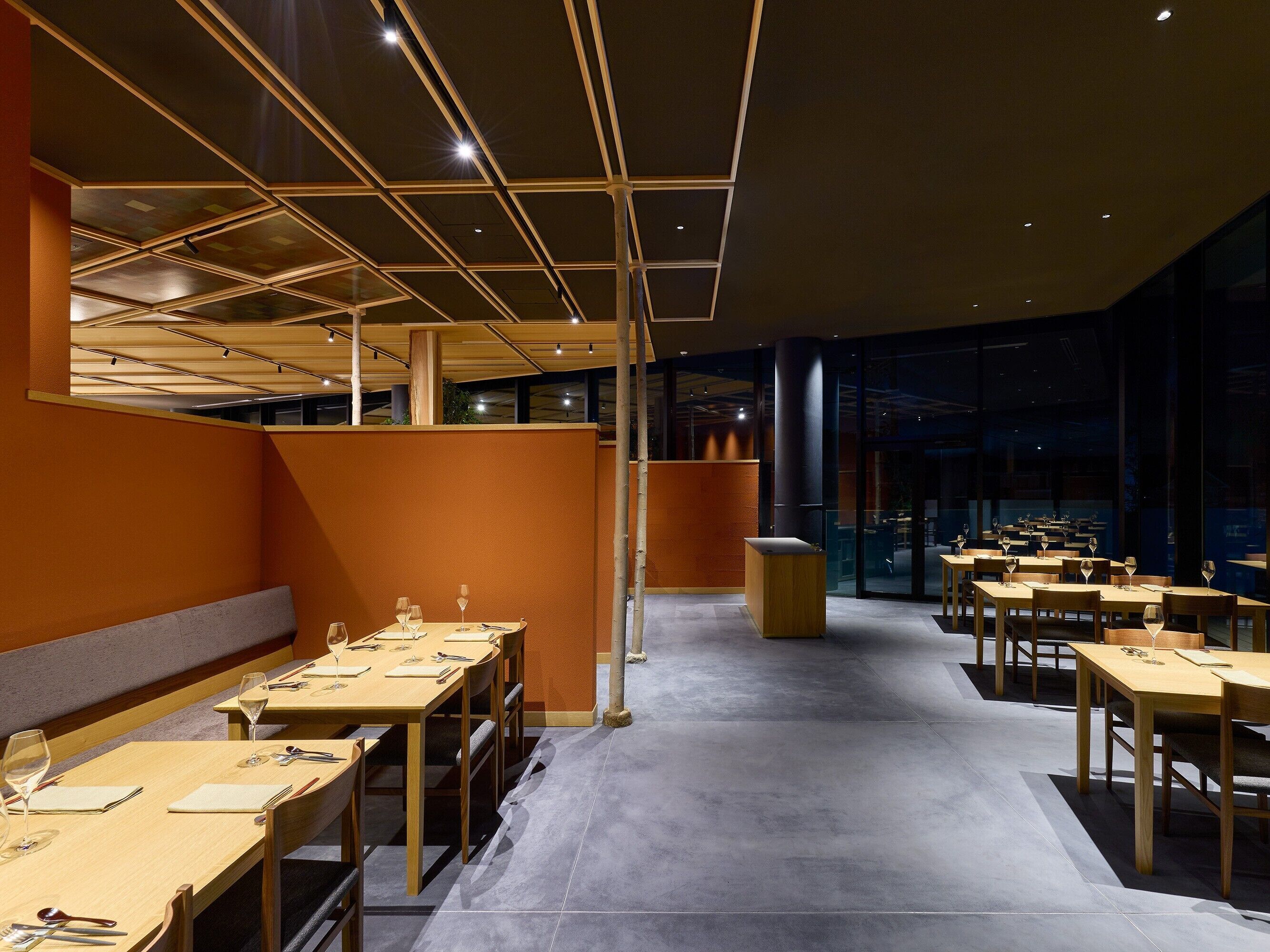 Ein modernes Restaurant mit Holztischen und -stühlen, einer zentralen Sitzgruppe und einer hohen Decke mit eingelassener Beleuchtung.