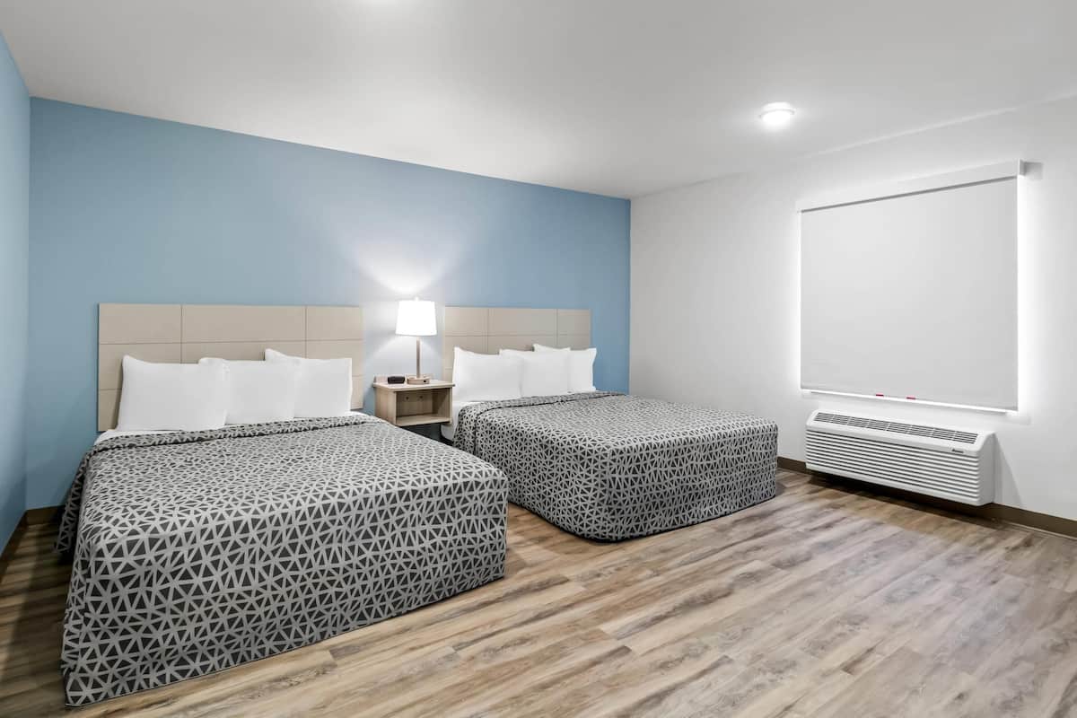 A hotel room with two beds, a nightstand, a lamp, and a wall-mounted air conditioner.
