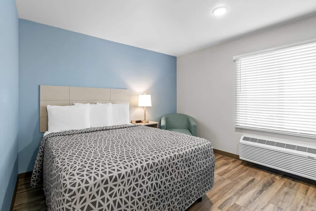 A hotel room with a bed, a nightstand, a lamp, a chair, and a window with blinds.