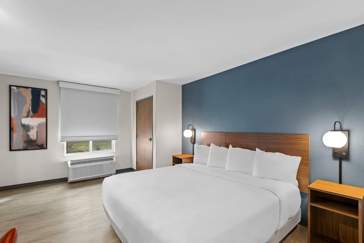 A modern hotel room with a large bed, two bedside tables, a nightstand, a window with blinds, a wall-mounted light, and a painting on the wall.