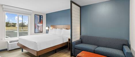 A modern hotel room with a large bed, a grey sofa, a small orange ottoman, and a window with a view of the outdoors.