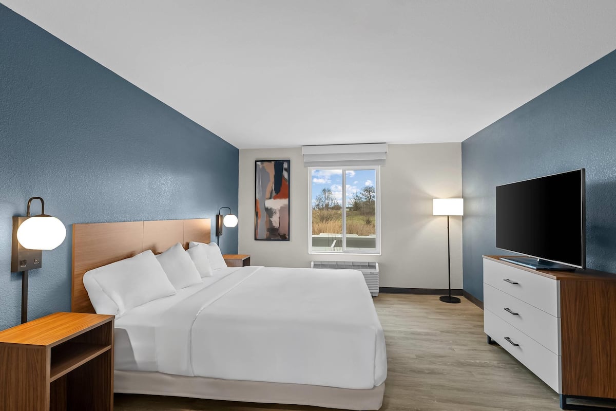 A modern hotel room with a large bed, bedside tables, a flat-screen TV, and a window with a view of the outdoors.