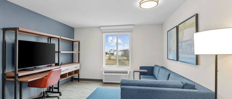 Een moderne woonkamer met een flatscreen televisie, een bureau met stoel, een blauwe bank en een raam met uitzicht op de omgeving.