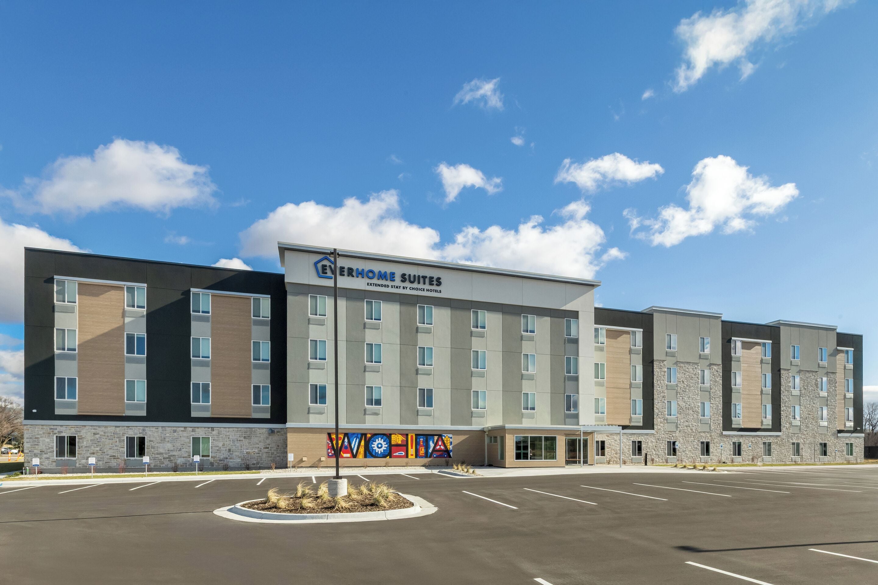 A modern hotel building with a sign reading "Everhome Suites" and a parking lot in front.