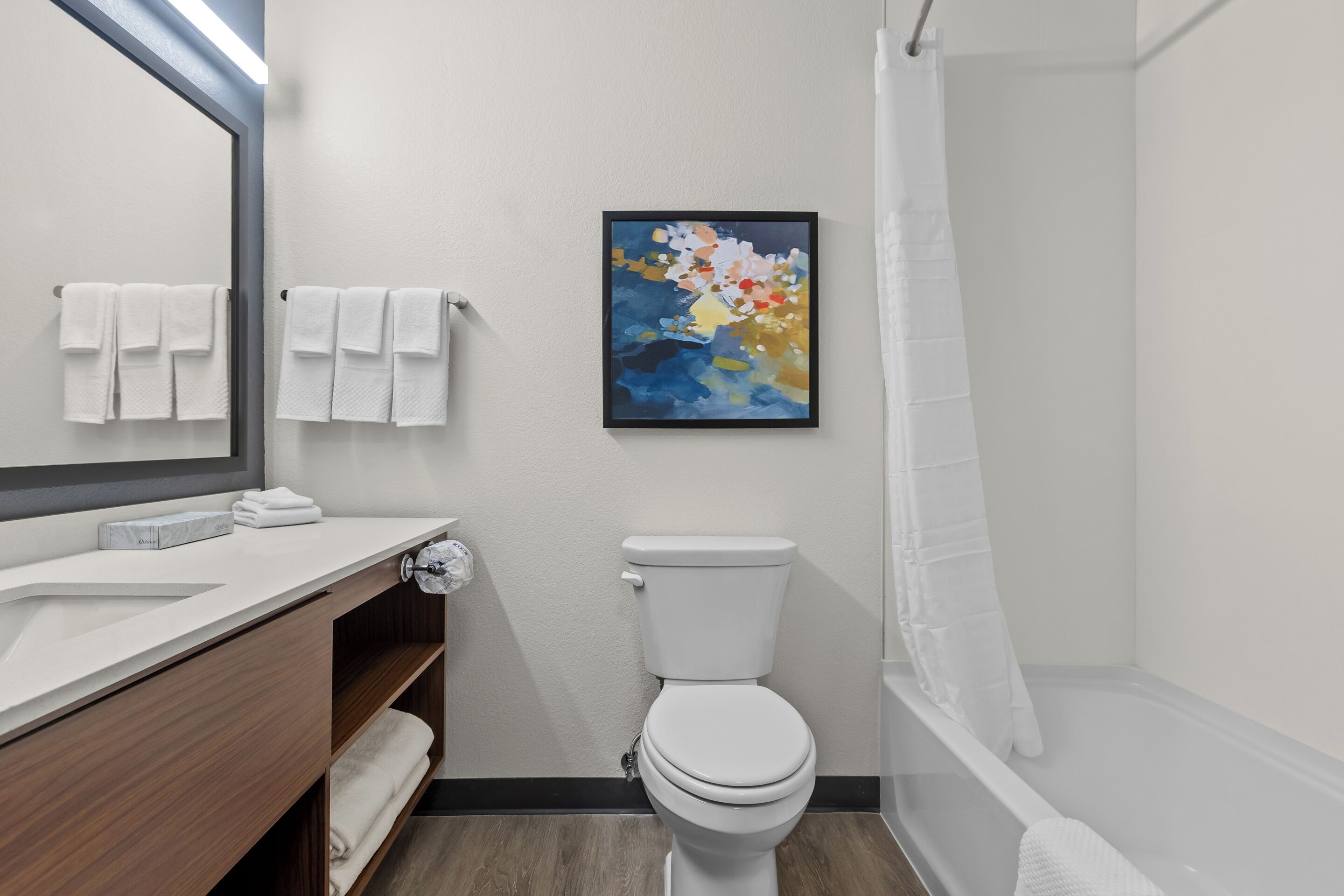 Une salle de bain moderne avec des toilettes blanches, un meuble-lavabo en bois doté d’un miroir et un tableau abstrait encadré accroché au mur.